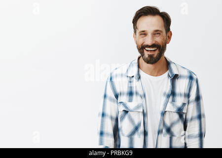 Gut aussehende glücklich und freundlich Mann mit Bart beiläufig Sprechen und Lachen mit Kollegen beim Mittagessen, stehend sorgenfrei über grauer Hintergrund, Spaß haben und sich mit interessanten Geschichten amüsiert Stockfoto