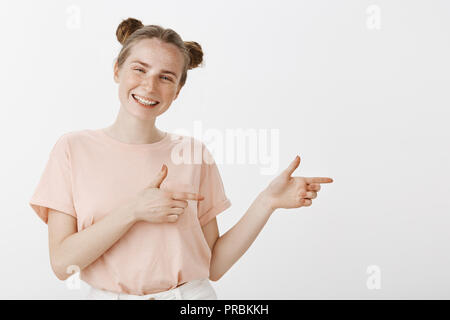 Studio geschossen von optimistisch gerne weibliche europäische Frau mit süßen Brötchen Frisur und Sommersprossen, nach rechts mit dem Finger gun Gesten, unbekümmert lächelnd, Richtung zeigen, toller Ort, um Spaß zu haben Stockfoto