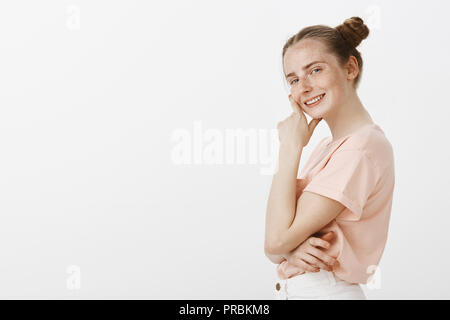 Mädchen will, um alle Details zu sehen. Neugierig gut aussehende trendy female mit Brötchen Frisur und Sommersprossen, stehend in Profil und schauen mit Interesse auf Kamera, die Hand in das Kinn, die fasziniert Stockfoto