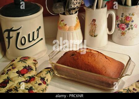 Frisch gebackenes Brot einer Banane Brot in einem Glas Container durch eine bunte Küche mitt, Mehl Topf und Geschirr, warmen Ambiente, Backen u. Kochen umgeben Stockfoto