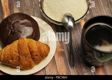 Eine Tasse schwarzen Kaffee, ein Teller mit Gebäck und eine Schüssel mit Zucker auf einem braunen Holzgrund Stockfoto