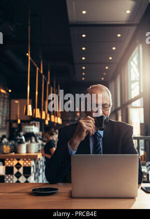 Vertikaler älterer Geschäftsmann am Laptop suchen und Kaffee im Cafe. Reifer Mann im Anzug im Cafe sitzen mit Laptop trinken Stockfoto