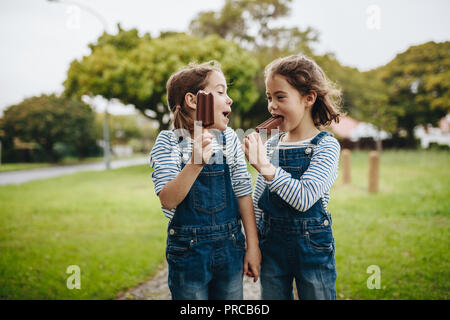 Zwei kleine Zwillingsschwestern in identischen Kleidung stehen im Freien und Schokolade essen Eis Süßigkeiten. Zwei kleine Mädchen genießen essen Süßigkeiten, Eis o Stockfoto