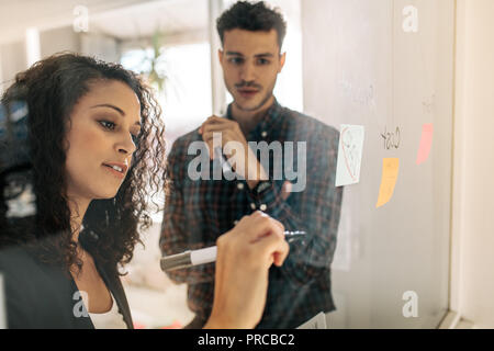 Bürokollegen diskutieren Geschäftsideen und -Pläne auf einer transparenten Glasplatte. Geschäftsfrau schreibt auf Haftnotizen auf transparentem Glas Stockfoto
