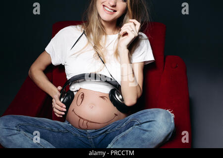 Junge schwangere Mutter und lächelnd Ohrhörer auf Ihrem Bauch. Stockfoto