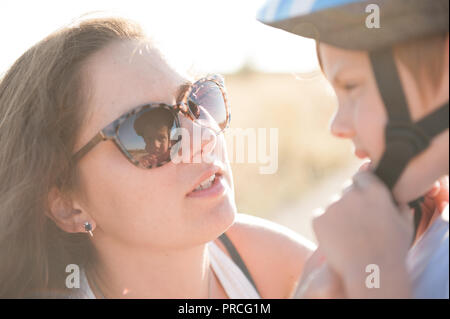 Junge schöne Mutter in trendigen Sonnenbrille hilft, Sohn sport Helm zu tragen Stockfoto