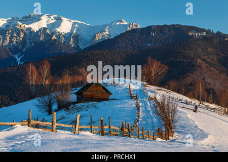 Alpine Landschaft mit rustikalen Holzmöbeln Cottage und schneebedeckten Grat der Bucegi-gebirge im Licht der untergehenden Sonne in einem kalten Winter Abend, in Kleie, Transyl Stockfoto