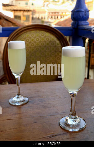 Zwei Glas der peruanischen Pisco Sour auf hölzernen Tisch mit einem Vintage Sessel und verschwommenes Gebäude im Hintergrund, Altstadt von Cusco, Peru Stockfoto