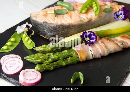 Gegrillter Lachs im Ofen gebacken, mit grünem Spargel im Speckmantel auf schwarzem Hintergrund gewickelt. Overhead. Stockfoto