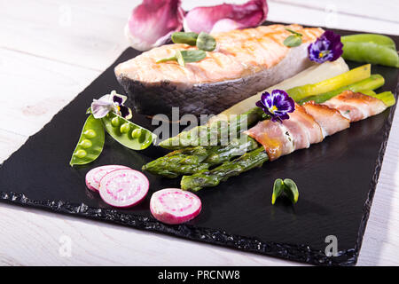 Gegrillter Lachs im Ofen gebacken, mit grünem Spargel im Speckmantel auf schwarzem Hintergrund gewickelt. Overhead. Stockfoto