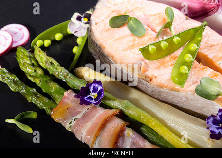 Gegrillter Lachs im Ofen gebacken, mit grünem Spargel im Speckmantel auf schwarzem Hintergrund gewickelt. Overhead. Stockfoto