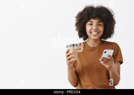 Taille-up Shot von niedlichen freundlich aussehende Städtische afroamerikanische Frau mit Afro Frisur holding Pappbecher mit Kaffee oder Tee und Smartphone in der Hand breit grinsend an Kamera news Lesen am Morgen Stockfoto