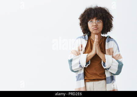 Taille- schuss unglücklich und düsteren miserabel afrikanische amerikanische Frau mit Afro Frisur in Gläsern schürzte die Lippen und finsteres Gefühl halten sie Hände und schaut in beten und beten zu Gott Stockfoto