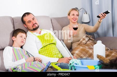 Müde Vater mit Sohn für Reinigung sitzen auf dem Sofa, während Mutter Fernsehen gekleidet Stockfoto