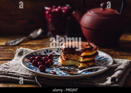 Quark Pfannkuchen mit Schokolade Topping, gefrorene Kirschen und Vanille Pod. Teekanne mit Löffeln und Glas von Beeren für den Hintergrund. Stockfoto