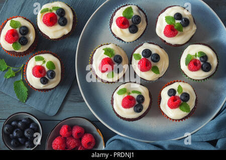 Chocolate Muffins oder kleine Kuchen mit Schlagsahne und Beeren, flach auf blauem Hintergrund legen Stockfoto