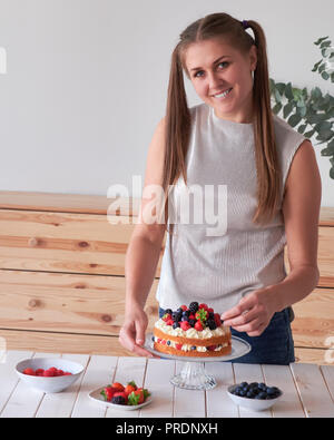 Junge lächelnde Frau in legere Kleidung Dekoration Kuchen mit Beeren lächeln in die Kamera Stockfoto