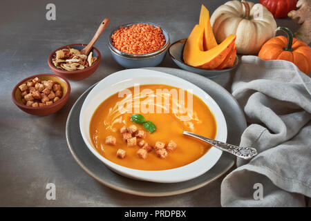 Kürbis und roten Linsen creme Suppe in Keramik Schüssel gewürzt mit Basilikum, Creme und Croutons, mit Zutaten und Gewürze um Stockfoto