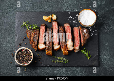 In Scheiben geschnittene Steak Roastbeef, gegrillt mit Pfeffer, Knoblauch, Salz und Thymian auf einer Schiefertafel Schneidebrett auf einem dunklen Stein serviert. Ansicht von oben Stockfoto