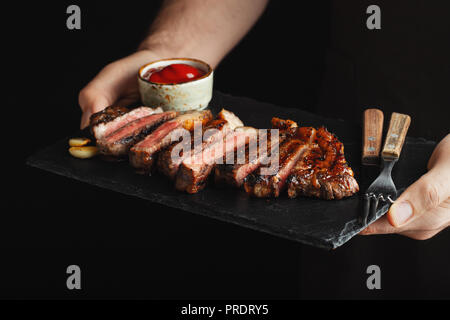 Mann, saftig gegrilltes Rindersteak mit Gewürzen und rote Sauce auf einem Stein Schneidbrett auf schwarzem Hintergrund Stockfoto