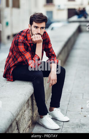 Junger bärtiger Mann, Modell von Mode, sitzen im städtischen Hintergrund legere Kleidung. Kerl mit Bart und moderne Frisur in der Straße mit Plaid Stockfoto