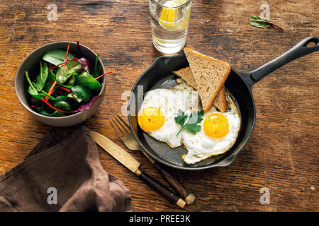 Frühstückstisch. Gebratenes Huhn Eier in der Pfanne mit Brot auf Holztisch mit Salat und Lemon Wasser serviert Stockfoto