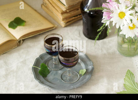 Creme de Cassis hausgemachte Schwarze Johannisbeere Likör in kleinen Gläsern, Bücher und Blumen. Im rustikalen Stil. Stockfoto