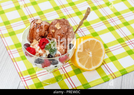 Schokolade und weißem Eis mit Himbeeren und Johannisbeeren, Beeren und Minze in einem Glas auf kariert Serviette und Zitrone auf hölzernen Schreibtisch Stockfoto