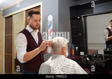 Mann mit grauen Haaren in Stuhl in Haarschnitt Kleid sitzen und sich die Haare schneiden. Professionelle friseur Pflege und Styling Haar des Menschen. Master tragen weiße Hemd und Weste. Stockfoto