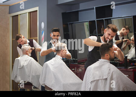 Prozess der drei professionelle Friseure arbeiten mit Kunden in Barbershop. Kunden tragen gestreifte Haarschnitt Kleider. Stattliche Friseure trimmen, Schneiden und Stylen Ihrer Haare. Stockfoto