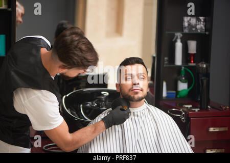 Professionelle männlichen Friseur in schwarz Handschuhe grooming Bart des Mannes mit der Bürste. Stattlicher Mann mit modischen Haarschnitt sitzen und Wegsehen. Konzept der Barbershop und Wellness. Stockfoto