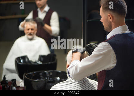 Professionelle Friseur in weißem Hemd, Weste und schwarzen Handschuhen Pflege und Styling Frisur seines Klienten. Reflexion im Spiegel der Mann und Meister. Konzept von Verfahren in der Barbershop und Haarpflege. Stockfoto