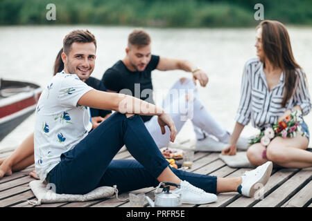 Hübscher junger Mann entspannt auf hölzernen Pier mit Freunden. Ferienhäuser Konzept Stockfoto