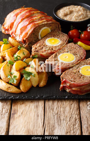 Köstliche geschnitten Meat Loaf in Speck mit Kartoffelecken und Sauce in der Nähe eingepackten auf einer Schiefertafel Board auf dem Tisch. Vertikale Stockfoto