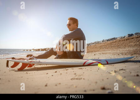 Reife männliche Surfer am Strand zu sitzen mit dem Surfbrett beobachten die Wellen, lens flare Stockfoto