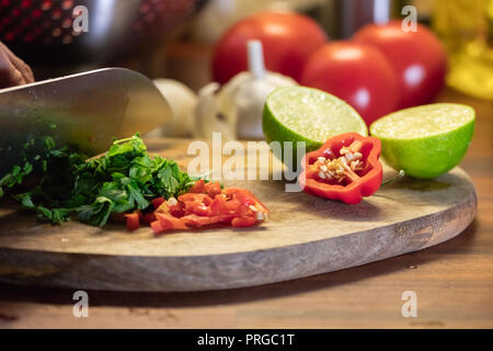 Kochen Tomaten Salsa-Hacken, Chilischote und Koriandergrün auf Holz Schneidebrett mit Knoblauch, Limette und Tomaten im Hintergrund Stockfoto