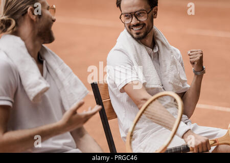 Lächelnd im Retrostil tennis Spieler sitzen auf Stühlen und Reden am Tennisplatz Stockfoto