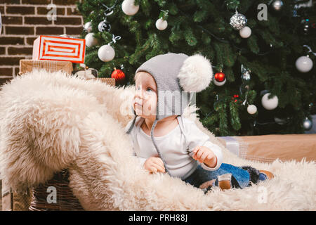 Betrifft Kinder Weihnachten neues Jahr. Kaukasische kleine lustige baby boy 1 Jahr alt sitzen Schlitten tragen Haut Weihnachtsbaum Kopf warme Mütze mit balabons Ba Stockfoto