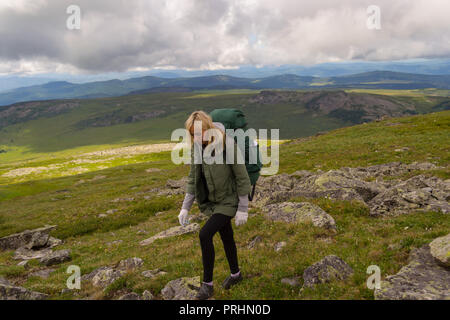 Junge schöne Frau reisen mit großen grünen Rucksack und Flasche, entlang der grünen Hügel mit grünem Gras zu Fuß auf den Felsen unter dem blauen Himmel und große Stockfoto