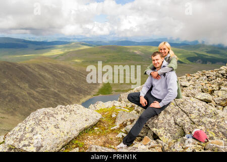 Der Kerl sitzt auf einem Felsen, sieht in die Kamera, hinter ihm ein blondes Mädchen lächeln vor dem Hintergrund der Wolken, blauer See, Tal von Hügeln. Eine liebevolle c Stockfoto