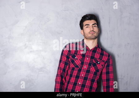 Eine ernsthafte hübscher junger Mann in einem rot kariertem Hemd stand vor einer grauen Wand in einem Studio. Stockfoto