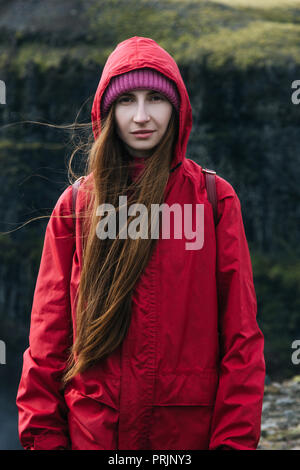 Attraktive junge Frau in Rot Regenmantel an Kamera mit Bergen im Hintergrund Stockfoto