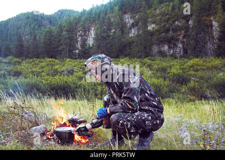 Der Mensch in der Tarnung Zubereitung von Essen am Lagerfeuer auf einer Lichtung in der Nähe der Bergen. der Stalker, Überleben im Wald Stockfoto