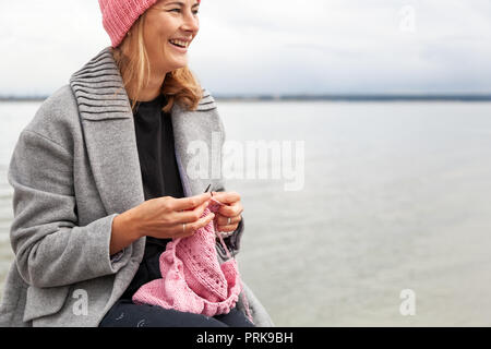 Close up og eine junge verheiratete Frau in modischen Mantel bekleidet Stricken einen rosa Hut mit Stricknadeln gegen Meer im sonnigen Herbsttag. Freiberufliche cr Stockfoto