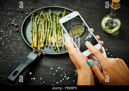 Die frau Hände halten das Telefon und ein Bild von köstlichen junge Spargel auf einem Holztisch Stockfoto