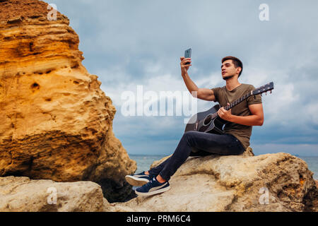 Junger Mann mit akustischer Gitarre bei Einnahme von selfie Phone an bewölkten Strand sitzt auf Felsen Stockfoto