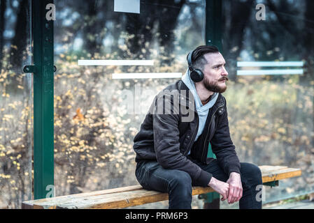 Ein hübscher junger hipster Mann an der Bushaltestelle warten, während das Hören von Musik auf seine Kopfhörer. Stockfoto