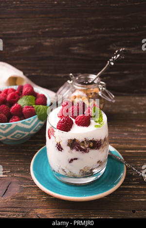 2-in-1-Joghurt mit Müsli und Himbeeren. In einem Glas, mit Minze dekoriert. Auf dunklen rustikalen Holztisch Hintergrund. Gesunde Ernährung Konzept. Stockfoto