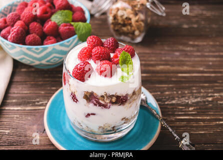 2-in-1-Joghurt mit Müsli und Himbeeren. In einem Glas, mit Minze dekoriert. Auf dunklen rustikalen Holztisch Hintergrund. Gesunde Ernährung Konzept. Stockfoto