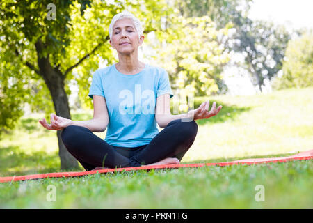 Ältere Frau im Lotussitz sitzen auf grünem Gras an summar Tag Stockfoto